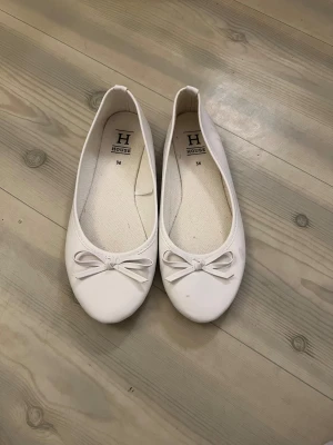 Vita ballerinaskor med rosett, House - Säljer ett par vita ballerinaskor från House i storlek 36. Skorna har rund tå, platt sula och en söt rosett framtill. Yttermaterialet är syntet och insidan är klädd i bomullstyg. Perfekta för dig som gillar enkel och stilren design.