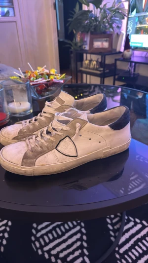 Philippe Model vita sneakers med grå detaljer - Säljer ett par Philippe Model Paris sneakers i vitt skinn med grå mockadetaljer och svart hälparti. Klassisk låg modell med snörning och rund tå. Snyggt sköldmärke på sidan och kontrastfärg på hälen. Perfekta för dig som gillar stilrena och exklusiva sneakers.