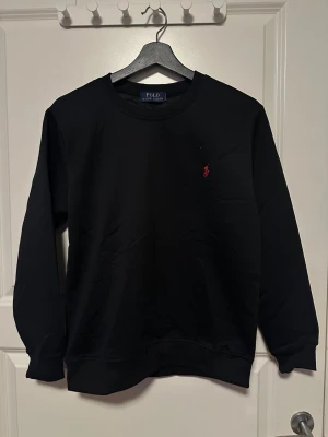 Svart sweatshirt från Polo Ralph Lauren - Svart sweatshirt från Polo Ralph Lauren med klassisk rund hals och lång ärm. Diskret röd Polo-logga broderad på bröstet. Mjuk och skön bomullskvalitet, perfekt för chill dagar. Stilren och enkel design som funkar till jeans eller joggers.