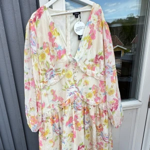 Plus blommig långklänning med volang från Boohoo - Romantisk långklänning från Boohoo i ljus bas med blommigt mönster i rosa, gul, grön och blå. Klänningen har v-ringning, långa ärmar, volangdetaljer och knappar framtill. Perfekt för sommarens tillfällen.