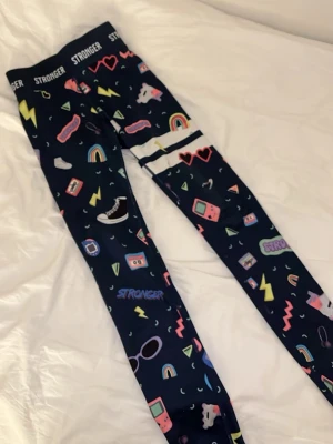 Mönstrade leggings från Stronger - Säljer ett par mörkblå leggings från Stronger med färgglada retro-motiv som regnbågar, blixtar, hjärtan och spelkonsoler. De har elastisk midja med logga och är gjorda i stretchigt syntetmaterial, perfekta för träning eller chill.