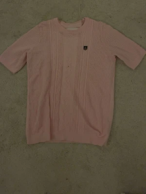 Rosa stickad tröja från Bondelid - Säljer en ljusrosa stickad tröja från Bondelid i storlek L. Tröjan har korta ärmar, rund hals och snygga kabelstickade detaljer framtill. På bröstet finns en liten patch med stjärna och ränder. Perfekt för vår och sommar! aldrig använd
