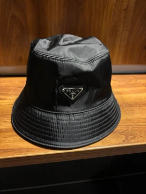 Svart bucket hat från Prada - Snygg svart bucket hat från Prada med ikonisk metalltriangel framtill. Hatten har en stilren och enkel design med glansigt syntetmaterial som ger en lyxig känsla. Perfekt accessoar för att lyfta din outfit och visa din stil.