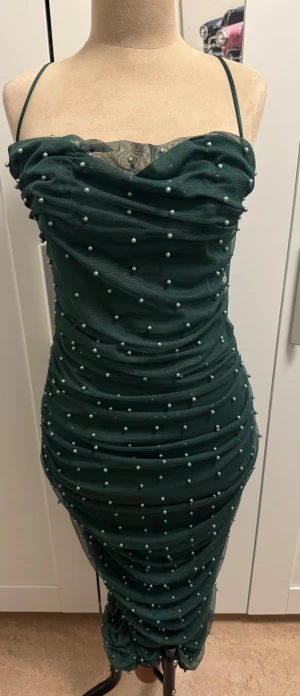 Mörkgrön pärlprydd klänning ASOS - Supersnygg mörkgrön klänning från ASOS DESIGN med smala axelband och tight passform. Klänningen är täckt av små pärlor och har draperad mesh över hela kroppen. Den är midilång och har dragkedja bak. Perfekt för dig som vill sticka ut med coola detaljer.