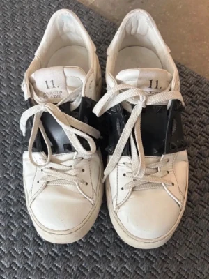 Valentino Rockstud Untitled sneakers - Säljer ett par Valentino Rockstud Untitled sneakers i vitt skinn med svart band och ikoniska pyramidnitar. Pris kan förhandlas