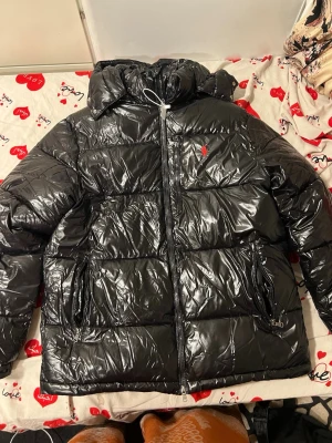 Svart pufferjacka från Polo Ralph Lauren - Säljer en svart, glansig pufferjacka från Polo Ralph Lauren med huva och dragkedjor på fickorna. Jackan har det klassiska röda Polo-logot på bröstet och är tillverkad i syntetmaterial. Perfekt för kalla dagar och har en riktigt modern vibe.