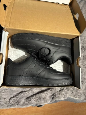 Nike Air Force 1 helsvarta sneakers - Säljer ett par klassiska Nike Air Force 1 sneakers i helsvart färg. Skorna har låg profil, rund tå och snörning. Tillverkade i skinn med en platt sula och diskret Nike-logga på sidan. Perfekta för dig som gillar stilrena och tidlösa sneakers.