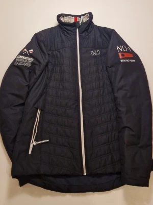 Mörkblå quiltad jacka Helly Hansen - Säljer en mörkblå quiltad jacka från Helly Hansen med vita detaljer och dragkedja. Jackan har broderade loggor och text på ärmarna, samt hög krage och praktiska fickor med dragkedja. Perfekt för dig som gillar segling och sportig stil.