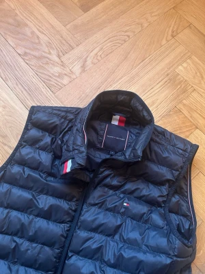 Svart dunväst från Tommy Hilfiger - Snygg svart dunväst från Tommy Hilfiger med quiltad design och hög krage. Västen har dragkedja framtill, två sidofickor med dragkedja och diskreta Hilfiger-detaljer i rött, vitt och blått på kragen och bröstet. Perfekt för lager-på-lager och chill stil.