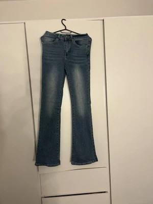 Blå bootcut jeans med broderade fickor - Snygga blå bootcut jeans med klassisk femficksdesign och coola broderade detaljer på bakfickorna. Jeansen har en lätt tvättad look och är tillverkade i mjukt denimtyg. Passar dig som gillar lite bredare ben och vill ha en unik touch med dekorerade fickor.