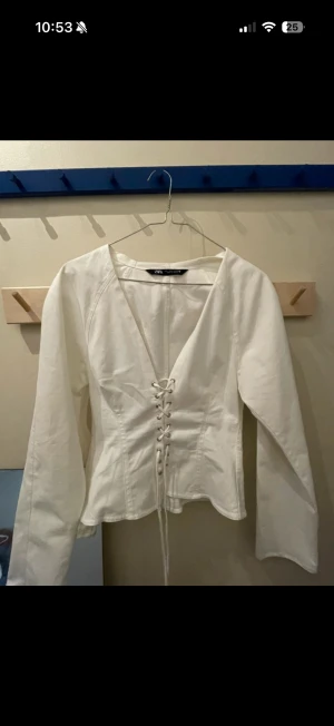 Vit korsettopp med snörning från Zara - Säljer en vit korsettopp från Zara med långärmad design och snygg snörning framtill. Toppen har v-ringad hals och figurnära passform som framhäver midjan. Materialet är bomull och toppen har en stilren look som passar till jeans eller kjol.