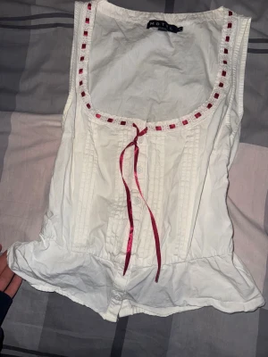 Vit linnetopp med rosett från Motel - Supersöt vit linnetopp från Motel med dekorativ röd rosett och band runt den fyrkantiga halsringningen. 