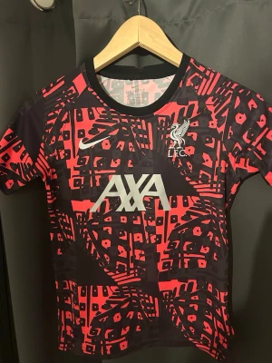 Nike Liverpool FC tränings t-shirt M - Säljer en Nike Liverpool FC tränings t-shirt i storlek M för barn med Dri-Fit material. Tröjan har ett grafiskt mönster i svart och rött, med vita loggor från Nike, AXA och LFC på bröstet. Rund hals och korta ärmar, perfekt för fotbollsträning.