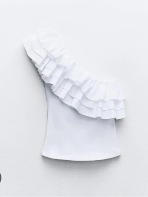 Vit oneshoulder topp med volang - Säljer en vit oneshoulder topp med flera lager volanger längs axeln. Toppens passform är tight och den är axelbandslös på ena sidan, vilket ger en snygg och trendig look. Perfekt för dig som vill sticka ut lite extra.