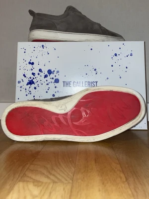 Christian Louboutin skor - Säljer ett par gråa sneakers från Christian Louboutin i mjuk mocka med klassisk röd sula och rund tå. Skorna har snörning och vit platt sula, samt diskreta sömmar och detaljer som ger en clean look. Perfekta för dig som gillar lyxiga och stilrena sneakers.