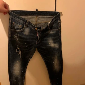 Mörkblå Dsquared2 jeans med färgstänk - Säljer ett par mörkblå jeans från Dsquared2 med coola slitningar och färgstänk i gult och vitt. Jeansen har klassisk femficksdesign, dragkedja och knapp, samt detaljerade sömmar. Perfekt för dig som gillar streetstyle och vill sticka ut.