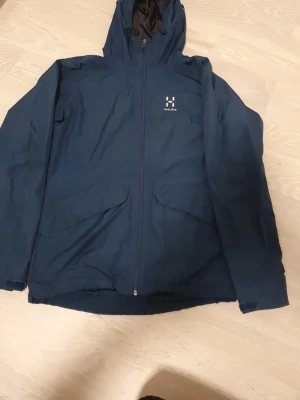 Blå vindjacka från Haglöfs med huva - Snygg marinblå vindjacka från Haglöfs med huva och hel dragkedja framtill. Jackan har två stora fickor med lock och ett diskret Haglöfs-tryck på bröstet. Insidan är fodrad med svart mesh för extra komfort. Perfekt för blåsiga dagar.