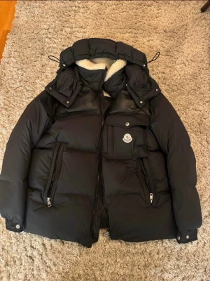Svart pufferjacka från Moncler - Säljer en svart pufferjacka från Moncler med stor huva och mysigt teddyfoder. Jackan har flera praktiska fickor med dragkedja och en bröstficka med Moncler-logga. Perfekt för kalla dagar med sin tjocka och fluffiga design. Stockholm, möts eller postar! 