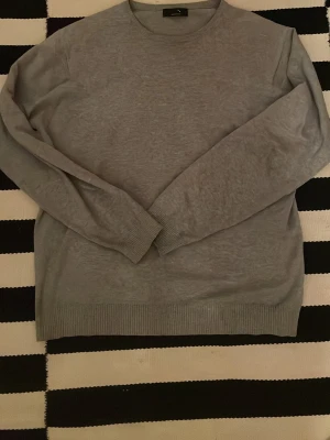 Grå stickad tröja från Selected - Snygg grå stickad tröja från Selected med rund hals och ribbade muddar vid ärmslut och nederkant. Perfekt för dig som gillar enkel och stilren design. Tröjan har lång ärm och är i mjukt material som känns skönt mot huden. Storlek L sitter som S-M