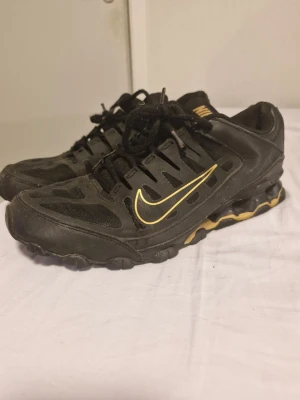 Svarta Nike sneakers med gul detalj - Nike sneakers i svart med snygga gula detaljer och logga på sidan. Skorna har snörning, rund tå och en robust platt sula med unikt mönster undertill. Ovandelen är i syntet och mesh för bra ventilation. Perfekt för dig som gillar sportig stil och streetwear.