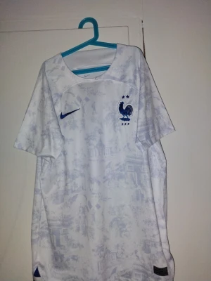 Frankrike tröja - Bilderna visar en vit Nike fotbollströja med blå detaljer och mönster, inte skor. Tröjan har Frankrikes landslagsmärke och Nike-logga, tillverkad i polyester med Dri-Fit-teknik. Tröjan har en liten svart fläck som knappt syns.