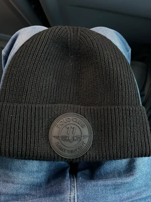 Svart stickad mössa Polo Club - Snygg svart stickad mössa från Polo Club Saint Vincent med ett stort runt gummimärke framtill. Mössan har bred uppvikt kant och är ribbstickad för extra stilpoäng. Perfekt för dig som vill ha en enkel men ändå trendig accessoar.