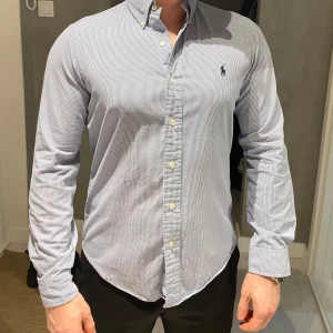 Blårandig skjorta från Ralph Lauren - Snygg blårandig skjorta från Ralph Lauren med klassisk button-down krage och broderad logga på bröstet. Skjortan är i smal passform och är tillverkad i mjuk bomull. 