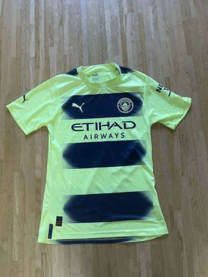 Manchester City gul/blå fotbollströja Puma - Säljer en Manchester City fotbollströja från Puma i neon-gul med mörkblå breda ränder och klubbmärke på bröstet. Tröjan har korta ärmar, rund hals och tryckta loggor. Perfekt för dig som älskar fotboll och vill sticka ut på planen.