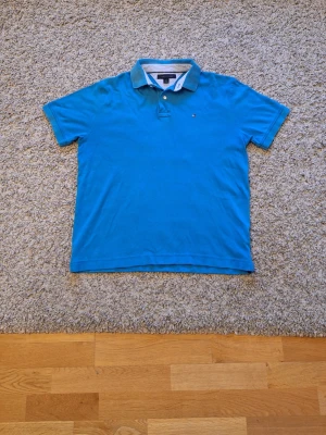 Blå Tommy Hilfiger pikétröja - Riktig snygg blå pikétröja från Tommy Hilfiger i storlek M. 100% bomull. Skicket är bra, inga slitningar eller defekter finns. Den har logga på vänstra bröstet och krage med två knappar. Passar riktigt bra med vad som helst. Skick: 8/10