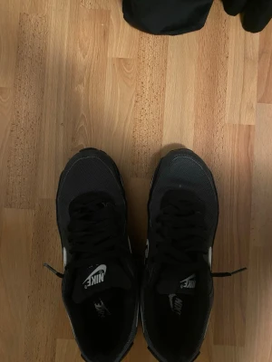 Svarta Nike Air Max sneakers - Säljer ett par svarta Nike Air Max sneakers med vit swoosh och detaljer. Skorna har mesh och syntetmaterial på ovansidan, samt snörning och platt sula. Perfekt för dig som gillar klassisk streetstil och vill ha bekväma sneakers med snygg design.