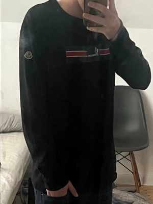 Svart långärmad topp från Moncler - Svart långärmad topp från Moncler med broderad logga på ärmen och ett tryckt mönster i blått, rött och vitt över bröstet. Rund halsringning och normal passform. Perfekt för dig som gillar stilrena designer med en touch av färg. Skriv för fler frågor