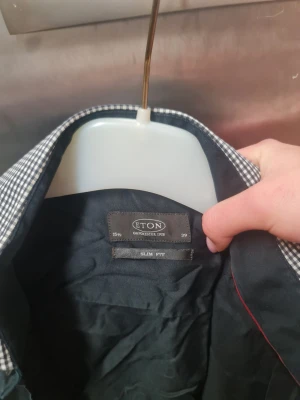 Svart slim fit skjorta från Eton - Svart skjorta från Eton i slim fit med klassisk krage och knappar framtill. Kragen och manschetterna har ett snyggt rutigt mönster i svart och vitt som ger en cool kontrast. Skjortan är tillverkad i bomull och har långa ärmar.