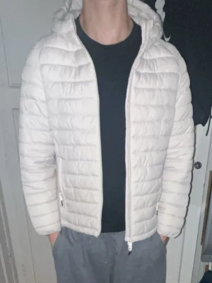 Vit pufferjacka med huva - Säljer en fräsch vit pufferjacka med huva och dragkedja framtill. Jackan har quiltad design med horisontella sömmar och två fickor med dragkedja på sidorna. Perfekt för kyliga dagar och har en clean look som passar till det mesta.