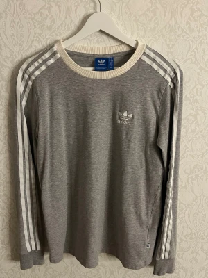 Grå långärmad tröja från Adidas  - Säljer en grå långärmad tröja från Adidas med klassiska vita ränder längs ärmarna och broderad logga på bröstet. Tröjan har ribbad vit krage och muddar, och är gjord i mjuk bomull. Perfekt för en sportig och chill stil.
