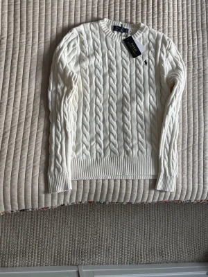 Vit kabelstickad tröja Polo Ralph Lauren - Vit kabelstickad tröja från Polo Ralph Lauren i storlek L. Tröjan har rund hals, ribbade muddar och det klassiska broderade Polo-logot på bröstet. Materialet är mjuk bomull och modellen är långärmad med normal passform.