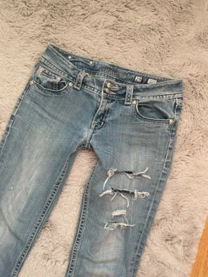 Miss Me ljusblå low waist bootcut jeans med slitningar - Säljer ett par ljusblå jeans från Miss Me med bootcut passform och snygga slitningar på ena benet. Jeansen har dekorativa bakfickor med broderade detaljer och pärlknappar. Klassisk femficksmodell i jeansmaterial med låg midja. Har sytt längst in i byxan men annars jättebra skick