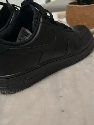 Svarta Nike Air Force 1 sneakers - Säljer ett par klassiska helsvarta Nike Air Force 1 sneakers med låg profil. Skorna har snörning, perforerad tå och platt sula. Tillverkade i skinn med svart Nike-logga på sidan och bak. Perfekt för dig som gillar stilrena och tidlösa sneakers.