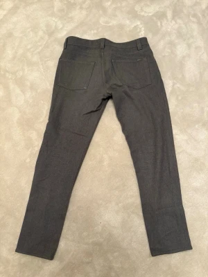 Grå kostymbyxor från Loro Piana - Snygga grå kostymbyxor från Loro Piana med klassisk rak passform och diskreta fickor bak. Byxorna har en stilren look med bälteshällor och knappstängning, perfekta för dig som gillar en clean och sofistikerad stil. Materialet känns mjukt och exklusivt. Nypris-9950kr/mitt pris-3000kr