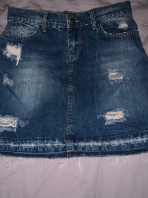 Blå jeanskjol med slitningar - Säljer en blå jeanskjol med coola slitningar och fransad nederkant. Kjolen har klassisk femficksdesign och knappstängning framtill. Perfekt för dig som gillar en avslappnad och trendig stil.