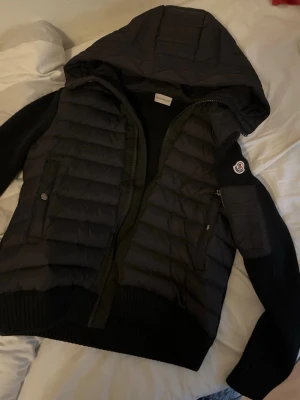 Svart Moncler Cardigan - Svart cardigab från Moncler med stickade ärmar och luva. Jackan har quiltad front, dragkedja och fickor med knappstängning. Moncler-logga på ärmen och snygga detaljer. Inget alls fel på jackan. Den är i storlek M, men dessa är lite små i storlek så passar ifall man vanligtvis har S. Den har NFC scan på loggan på armen och qr kod inuti.