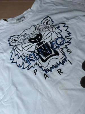 Vit Kenzo t-shirt med tigertryck - Säljer en vit t-shirt från Kenzo med ikoniskt tigertryck och glittriga blå detaljer på bröstet. T-shirten har rund hals och korta ärmar, tillverkad i mjuk bomull. Perfekt för dig som vill sticka ut med ett coolt statement-plagg.