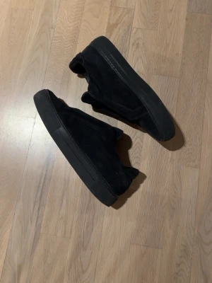 Axel Arigato - Säljer ett par svarta sneakers med platt sula och rund tå. Skorna är tillverkade i mocka och har en stilren, minimalistisk design utan synlig snörning. Perfekta för dig som gillar en clean look och vill ha något som funkar till många olika outfits.