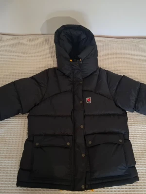 Svart Fjällräven Expedition dunjacka Dam som ny - Säljer en svart Fjällräven Expedition Down Lite Jacket med huva och gula detaljer. Jackan har två stora fickor med knappar, dragkedja och tryckknappar framtill. Fylld med dun och har ett klassiskt Fjällräven-märke på bröstet. Perfekt för kalla dagar. Storlek Xl dam men passar Large med!   Rek pris 7499kr Inköpt förra säsong av mig använd några ggr. Så nära ny/butik skick man kan komma. 