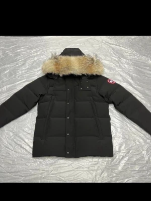 Canada Goose jacka  - Svart pufferjacka från Canada Goose med stor huva och beige pälsdetalj. Jackan har flera fickor framtill, knappar och dragkedja. Klassisk logga på ärmen och quiltad design som ger extra värme. Perfekt för kalla vinterdagar.