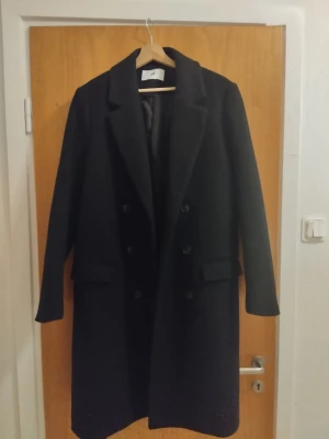 Svart ullkappa i storlek 44 - Svart lång kappa från H&M med klassisk krage och dubbelknäppning framtill. Kappan har två fickor med lock och ett bälte bak för extra stilpoäng. Materialet är ullblandning med 66 procent ull. Den är alltså väldigt varm.. Perfekt för dig som gillar clean och tidlös look. Kappan är i nyskick.