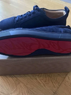 Mörkblå sneakers från Christian Louboutin - Säljer ett par mörkblå sneakers från Christian Louboutin med klassisk röd sula. Skorna har snörning, rund tå och är tillverkade i mocka med beige innersida. Perfekt för dig som vill ha en lyxig och stilren look med exklusiv känsla.