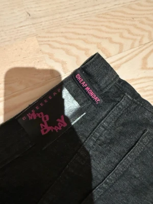 Svarta jeans från Cheap Monday - Säljer ett par svarta jeans från Cheap Monday med klassisk femficksdesign och breda ben. Jeansen har en mörk tvätt och är tillverkade i jeansmaterial med en cool etikett i rosa. Perfekt för dig som gillar avslappnad stil och vill ha lite extra attityd i garderoben. Storlek 32/34 endast använda vid ett tillfälle!