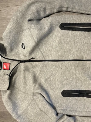 Nike tech hoodie - Säljer en stilren och bekväm Nike Tech hoodie i grå färg, storlek M. Hoodien är tillverkad i Nike Tech Fleece-material som ger bra värme utan att kännas tung. Modern passform med dragkedja, huva och praktiska fickor. Passar perfekt till både vardag och träning.  Skick: Bra skick Storlek: M Färg: Grå Märke: Nike
