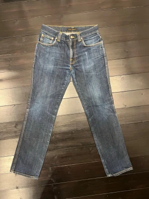 Nudie Jeans Slim Jim Dry Japan W31 L32 - Säljer ett par Nudie Jeans Slim Jim Dry Japan i klassisk blå tvätt med snygga kontrastsömmar och broderade bakfickor. Modellen har normal passform och raka ben, tillverkade i 100% bomull. Perfekt för dig som gillar stilrena jeans med coola detaljer.