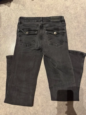 Svarta bootcut jeans 34 - Svarta jeans med bootcut-snitt och klassisk femficksdesign. Snygga detaljer med ficklock och silverfärgade knappar bak. Jeansen har normal passform och är tillverkade i jeansmaterial med lätt tvättad look. Perfekt för dig som gillar en lite bredare benslut.                                                                             Mått: 	•	Midjemått: ca 34 cm 	•	Grenhöjd: ca 21 cm 	•	Innerbenslängd: ca 74 cm